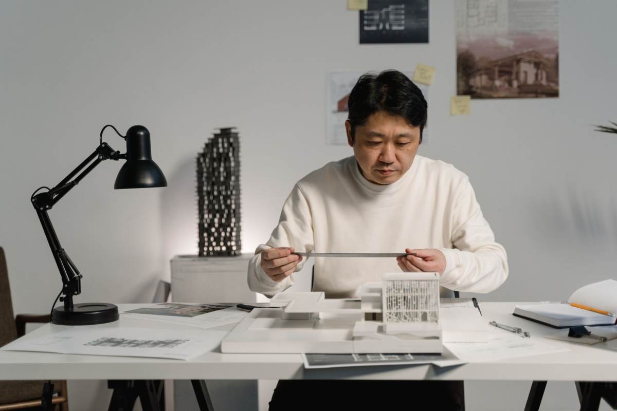 An architect at a white desk measuring a small architectural model beside a desk lamp