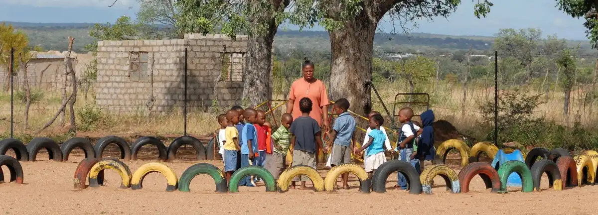 A teacher stands with a circle of young children holding hands in a playground edged with painted yellow, green and red tyres