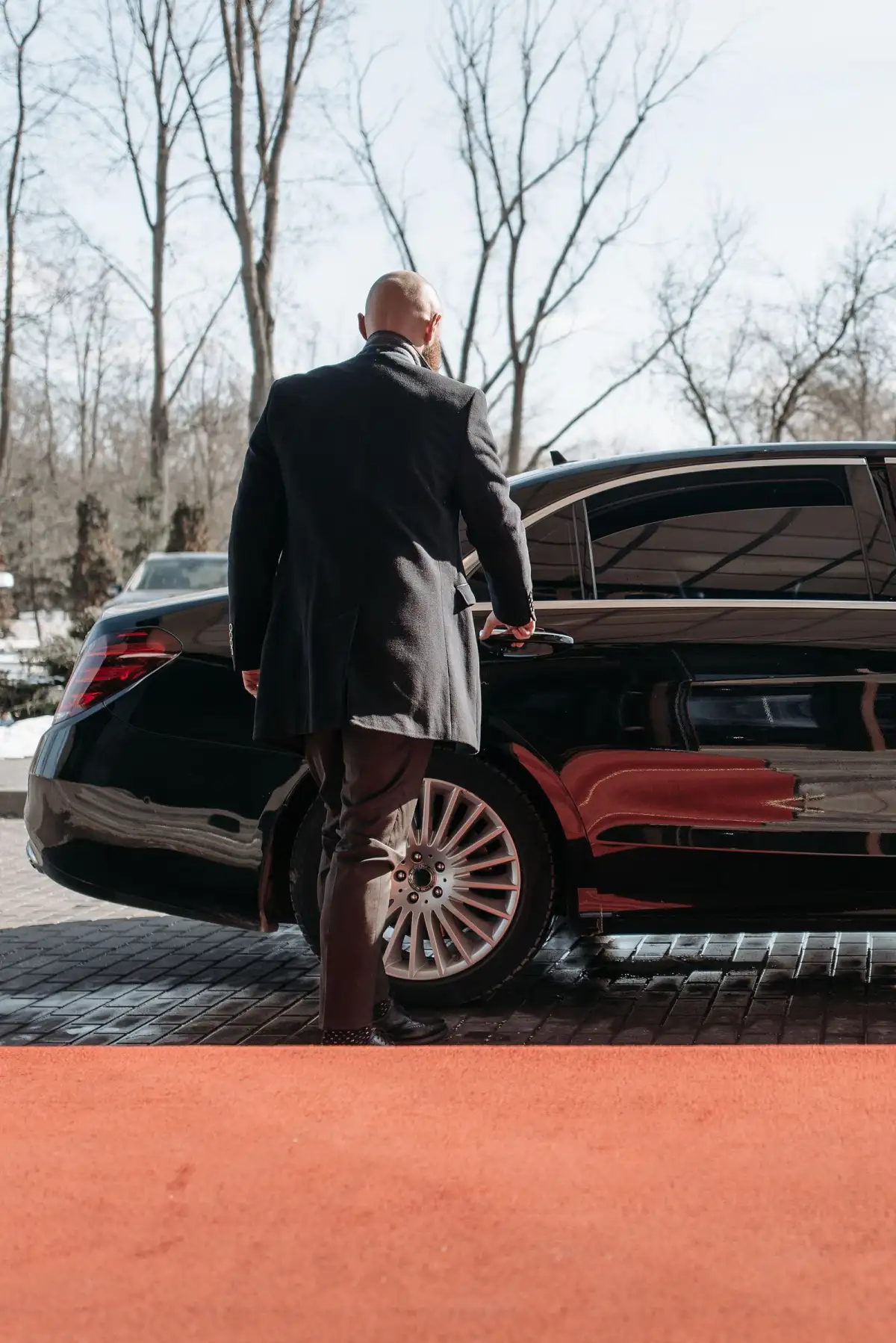 A chauffeur in a long dark coat opens the rear door of a black saloon car on a paved forecourt