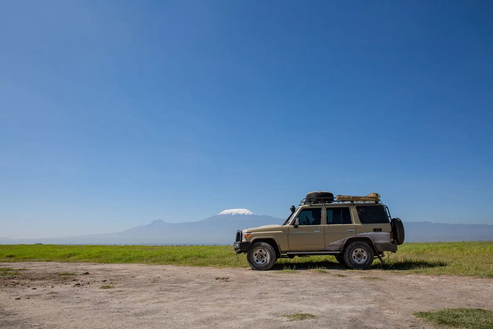 A tan safari 4x4 with a roof rack and spare tyre parked on short grass, with the snow-capped peak of Mount Kilimanjaro behind it under a deep blue sky