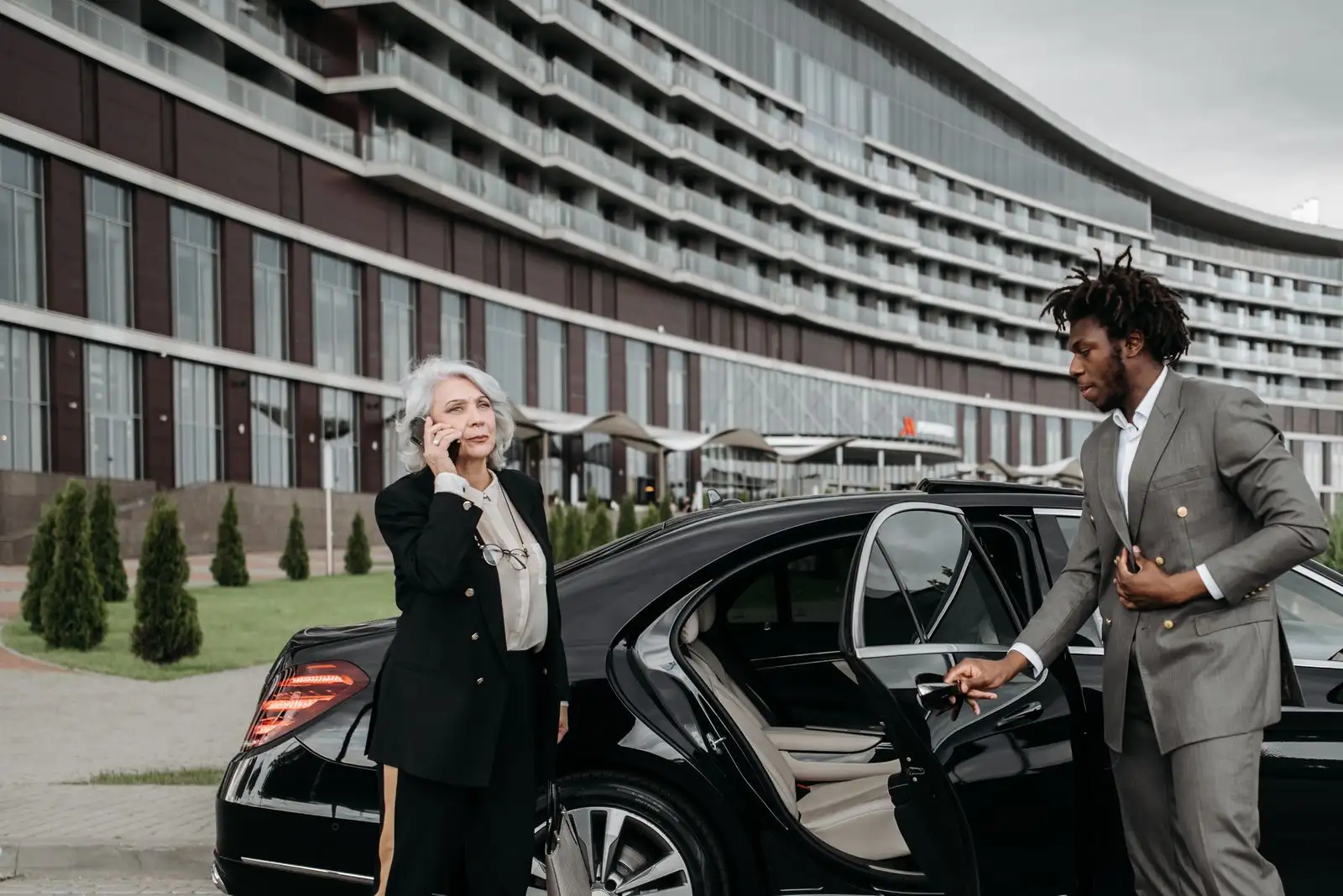 A chauffeur in a grey suit holds the rear door of a black saloon car for a woman on the phone outside a large hotel