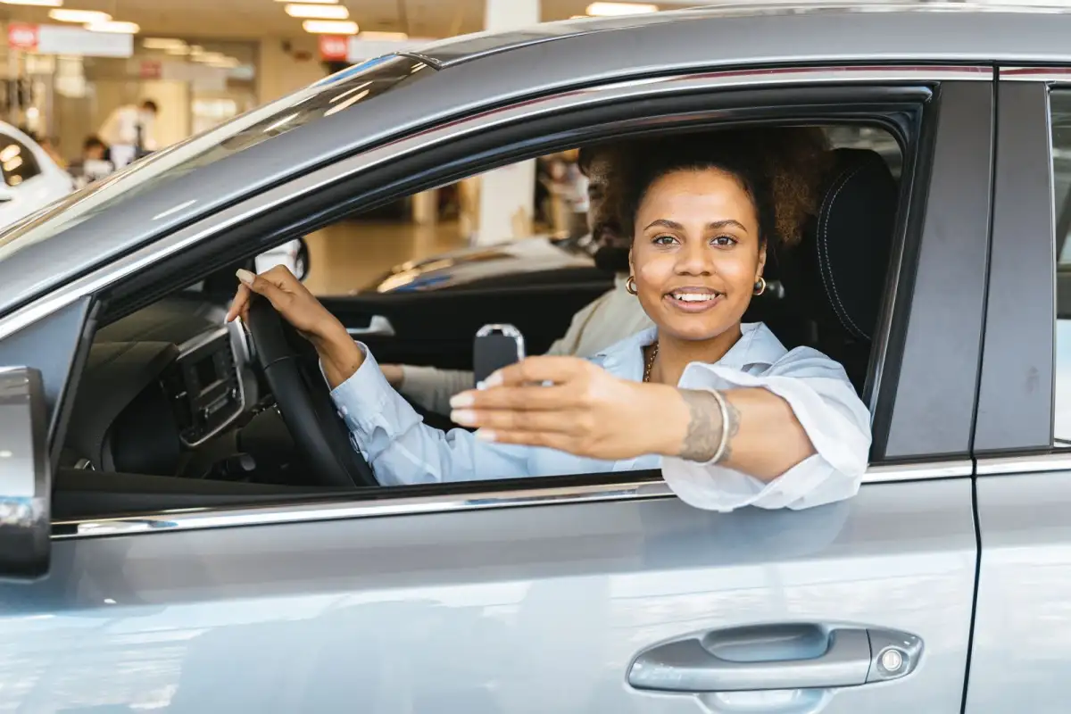 A smiling woman at the wheel of a silver car holds a key fob out of the open window