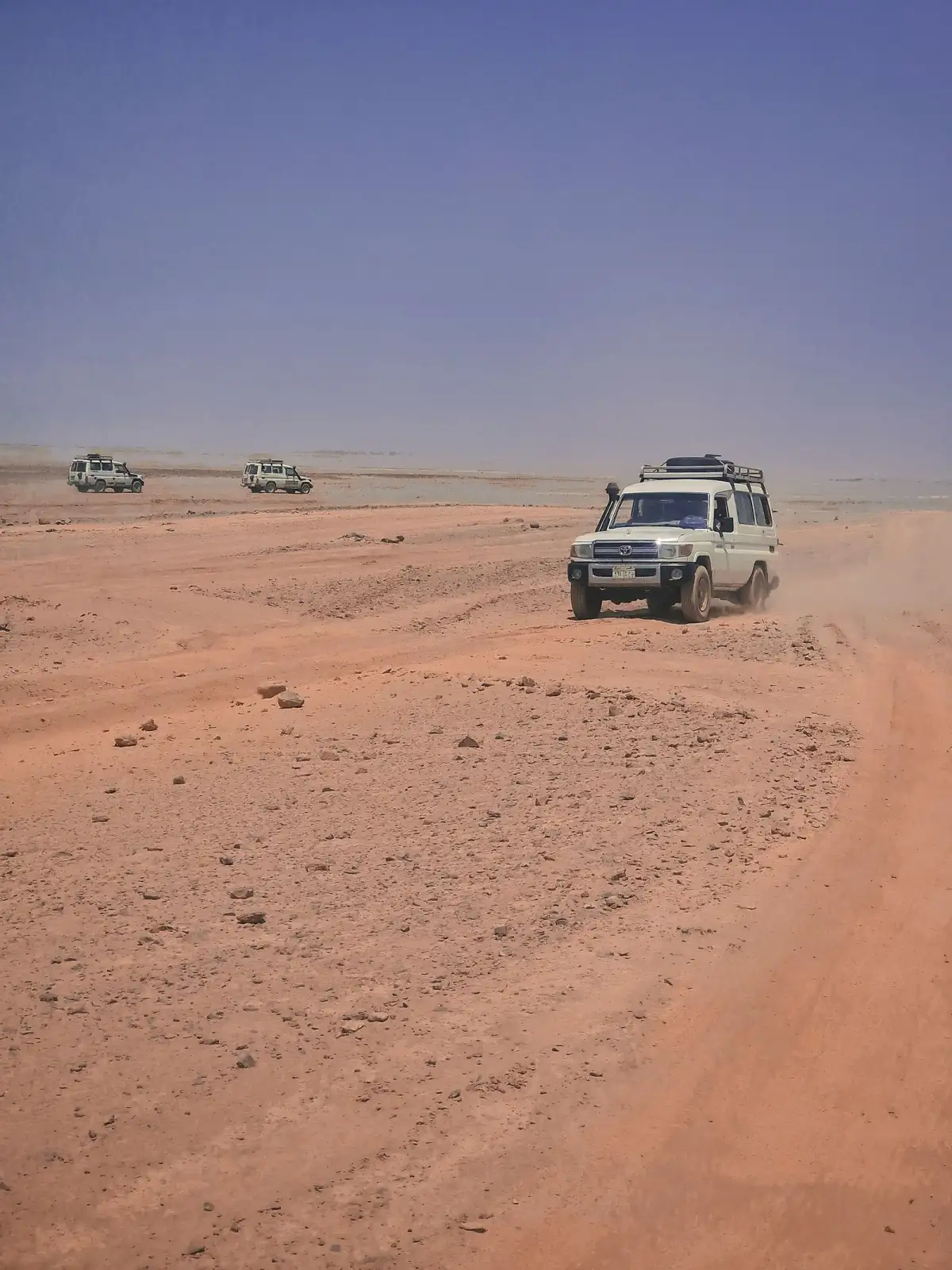 Three safari 4x4s drive in a line across a wide, dry, red-orange plain under a hazy sky