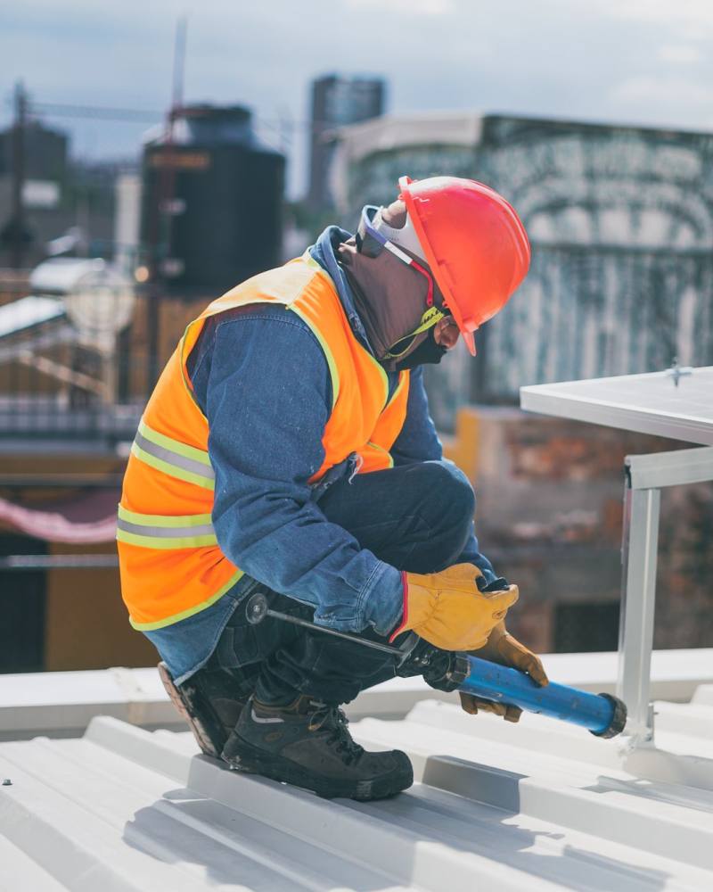 A technician in a hard hat and hi-vis vest sealing a mounting bracket on a corrugated roof beside a solar panel