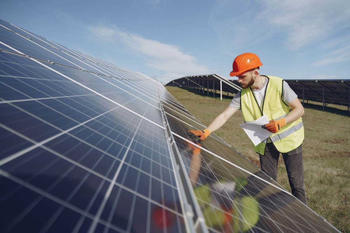 An engineer in a hard hat and hi-vis vest inspecting solar panels with paperwork in hand