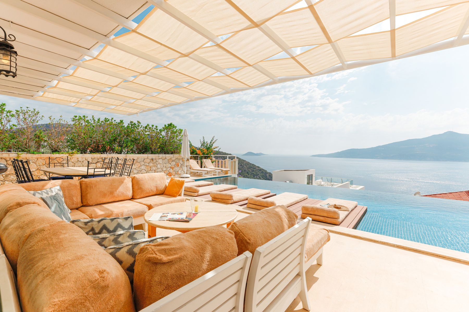 A sunlit hotel terrace with lounge seating beside an infinity pool overlooking the sea