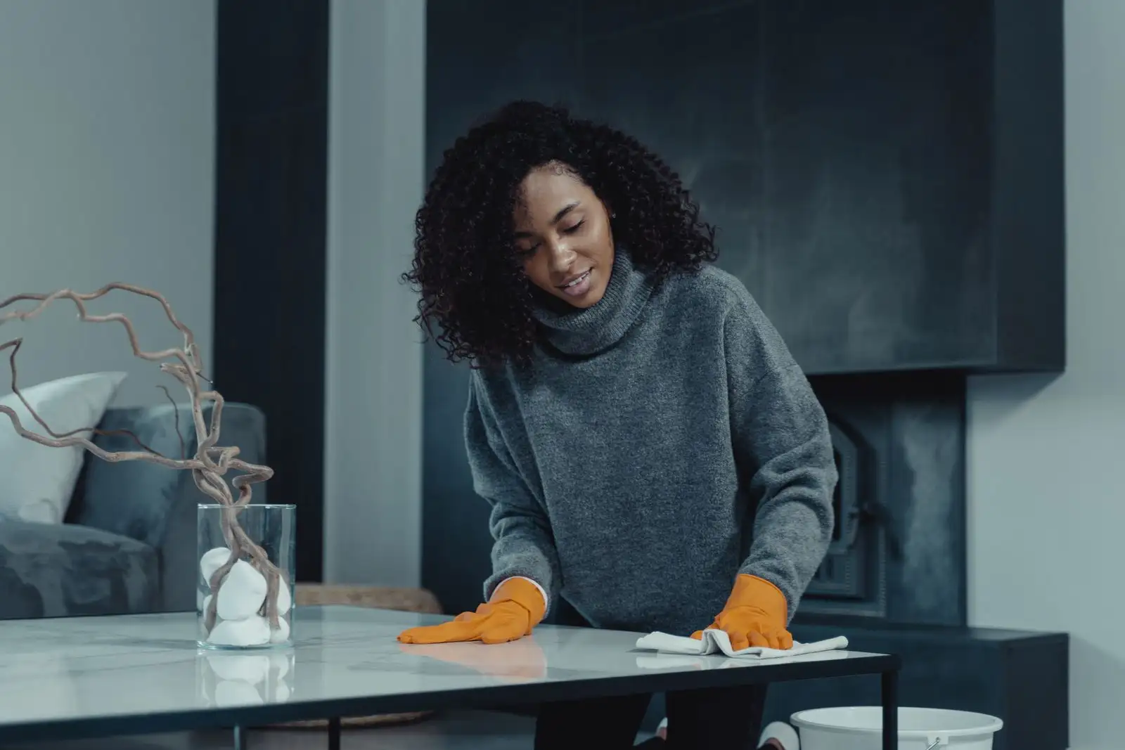 A woman in a grey jumper and orange gloves wipes a white marble coffee table in a bright, modern living room