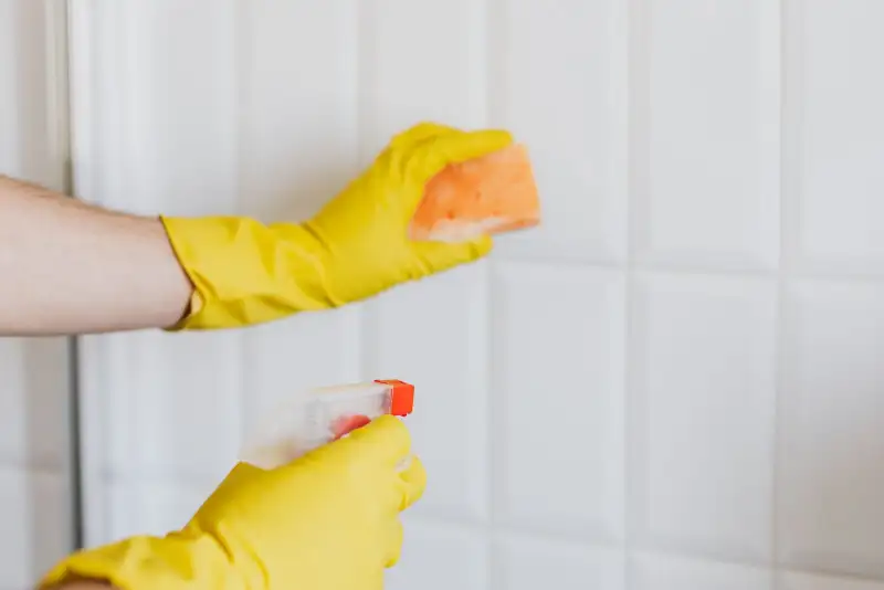 Two hands in bright yellow rubber gloves hold a sponge and a spray bottle in front of white wall tiles