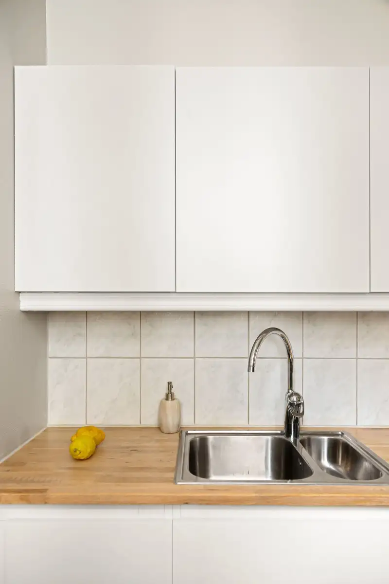 A bright empty kitchen with white cupboards, a stainless steel sink and a wooden worktop