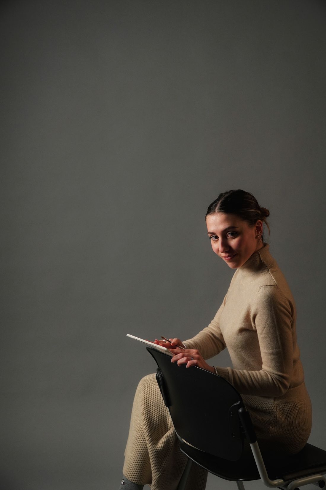 A woman in a beige outfit seated on a chair holding a tablet against a grey studio backdrop